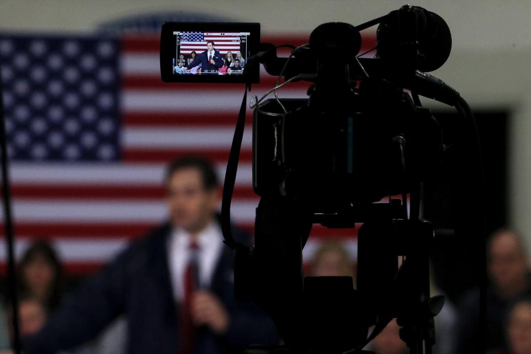photograph of a camcorder filming a white man in a suit speaking in front of a US flag